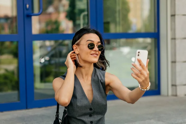 Gadis cantik yang menarik dengan gaya rambut pendek memakai kacamata hitam membuat selfie di kota sambil beristirahat di akhir pekan — Stok Foto