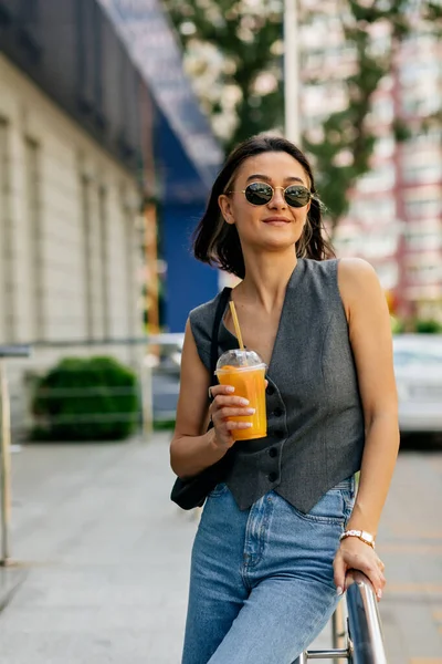 Fancy girl with short hairstyle wearing sunglasses and vest holding orange summer drink and smiling while walking in the city — 스톡 사진