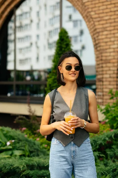 Stylish tanned girl in sunglasses holding summer drink. Outdoor portrait of debonair laughing woman walking on background of summer cafe — 스톡 사진