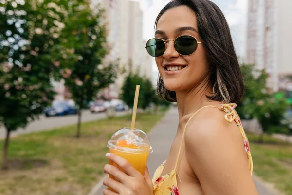 Close up portrait of lovely smiling young woman in sunglasses with orange juice turn around at camera on city background — 스톡 사진