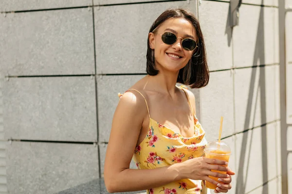 Wanita cantik bergaya dalam gaun musim panas dan kacamata hitam menatap kamera dengan senyum bahagia saat minum jus di luar — Stok Foto