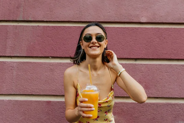 Detail portrét usměvavé stylové dívky s krátkými vlasy na sobě letní šaty dotýkají její vlasy a drží pomerančovou šťávu přes růžové pozadí — Stock fotografie