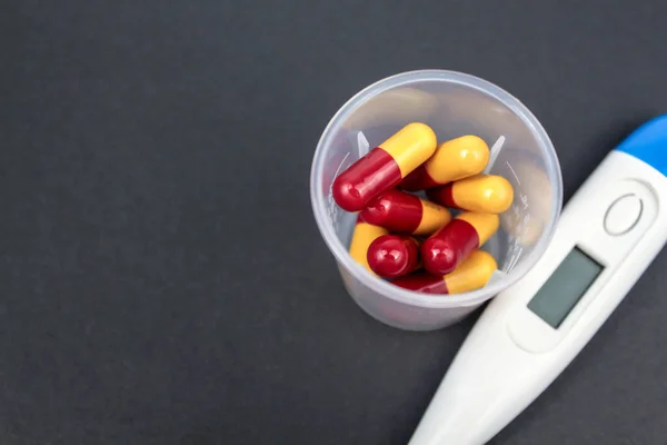 Medical capsules, tablets and clinical thermometer inside measuring cup on black background. Side view