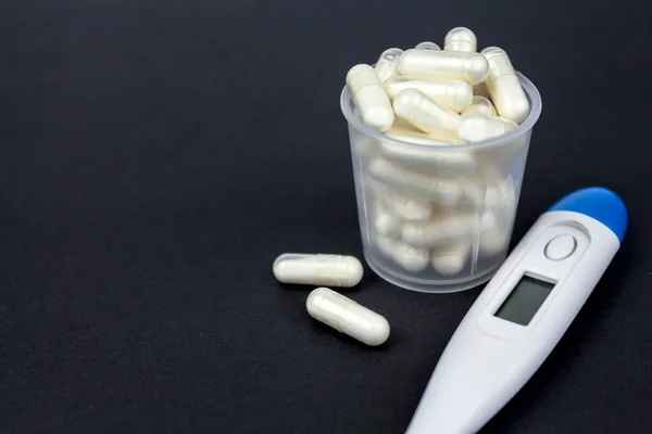 Medical capsules, tablets and clinical thermometer inside measuring cup on black background. Side view