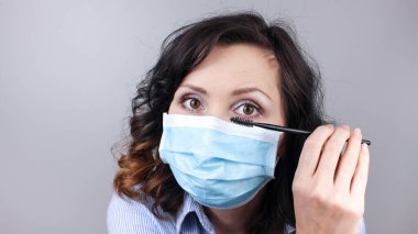Koronavirüse karşı koruyucu maske takan ve makyaj yapan bir kadın. Maskeli bir kadın aynaya bakıyor ve rimel sürüyor. Tıbbi maske, yakın çekim, Covid19 önleme