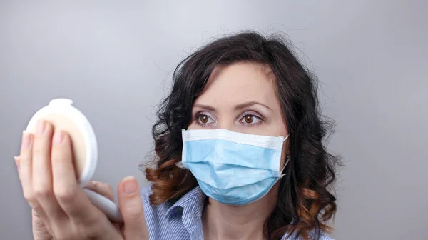 Woman wearing protection face mask against coronavirus and applying make-up. Woman in a mask looking in a mirror. Medical mask, Close up shot, Select focus, Prevention from covid19