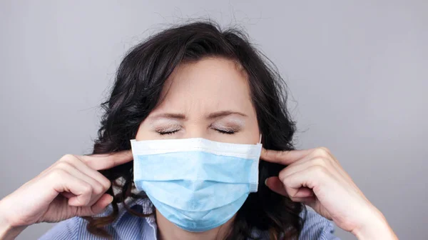 Portrait of nervous brunette caucasian young woman with surgical medical mask standing and putting finger on ears and don\'t want to hear. studio shot, isolated on grey background.
