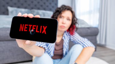 Ottawa ON, Kanada - Ekim 06, 2025: Kıvırcık saçlı kadın Netflix logolu akıllı telefonu modern oturma odasında yerde otururken gösteriyor, yükleme sorunundan duyduğu hayal kırıklığını dile getiriyor