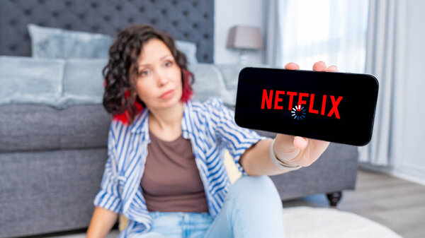 Оттава ON, Канада - 06 октября 2025 года: Женщина в повседневной одежде сидит на полу, показывая смартфон с экраном загрузки Netflix, иллюстрируя потоковые трудности в современном жилом пространстве