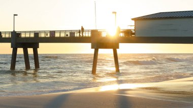 İnsanlar gün batımında bir rıhtımda yürüyorlar, dalgalar kumlu sahilde süzülüyor, okyanus kıyısında sakin bir anı yakalıyorlar.