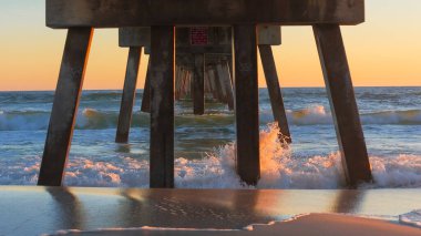 İskele yapısı, gün batımında düşen okyanus dalgalarının üzerinde yükselir. Islak kumu yansıtır ve renkli gökyüzü ile sakin bir sahne yaratır.