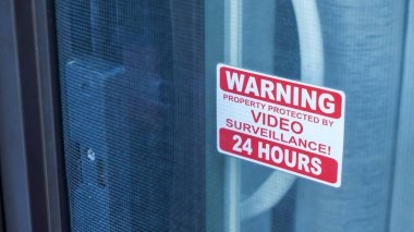 Uyarı levhası, güvenlik özellikleri ve güvenlik önlemlerinin altı çizilerek kapının üzerinde açıkça görülen güvenlik kameraları tarafından izlendiğini bildirir.