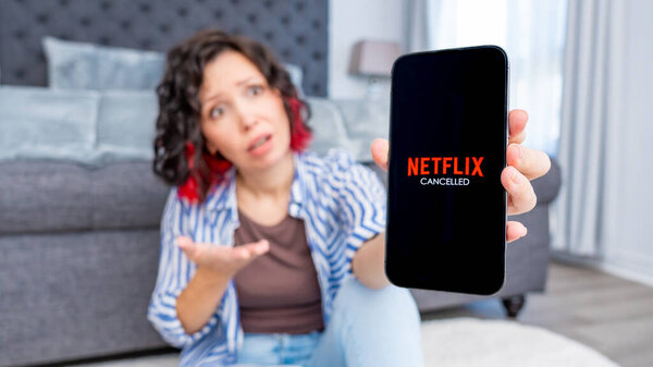 Оттава ON, Канада - 06 октября 2025: Кудрявая женщина держит смартфон, показывающий Netflix отмененное сообщение, выглядящее разочарованным в стильной гостиной с уютной атмосферой