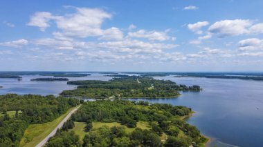 Aerial perspective of a tranquil lake bordered by vibrant green trees and islands, with a clear blue sky and soft clouds enhancing the peaceful atmosphere