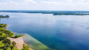Berrak mavi sulara, kumlu plajlara ve canlı yeşilliklere sahip sakin göl perspektifi, barışçıl bir açık hava ortamı yaratıyor.