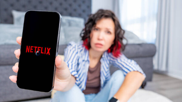 Оттава ON, Канада - 06 октября 2025: Кавказская женщина показывает смартфон с логотипом Netflix, сидя на полу в стильной гостиной, выражая разочарование по поводу проблем с приложениями