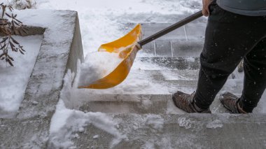 Şahıs, kar fırtınası sırasında dış basamaklardan kar temizliyor, kış koşullarını vurguluyor ve karların temizlenmesi için gereken fiziksel çabayı gösteriyor