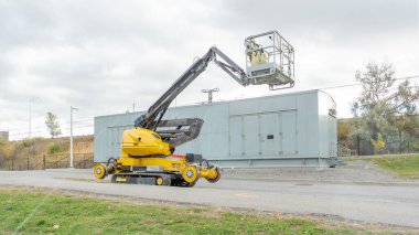 Sarı hava asansörü bir inşaat sahasında, çalışanları yükselterek, bulutlu bir arkaplanı olan büyük bir sanayi binasının yakınındaki bakım işleri için