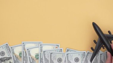 El, siyah bir uçak oyuncağıyla para yığınlarının üzerine konmuş durumda. Seyahat planlama ve finansal hedefler kavramını gözler önüne seriyor. Kamera onu yukarı doğru takip ediyor.