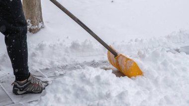 Şahıs, karlı bir manzarayla çevrili sarı bir kürekle aktif olarak kaldırımdan kar kürüyor ve kış mevsimleri ve çabalarını vurguluyor.