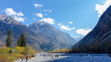 Bişkek, Kırgızistan - 29 Eylül 2024: Ala-Archa Doğa Parkı 'nın görkemli dağlarla ve renkli sonbahar ağaçlarıyla çevrili kayalık nehir yatağındaki yürüyüşçülerin yer aldığı manzara