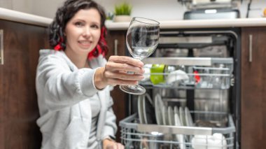 Kısa siyah saçlı bir kadın, çağdaş bir mutfakta temiz bulaşıklarla dolu açık bir bulaşık makinesinin yanında dururken temiz bir bardak sunar.