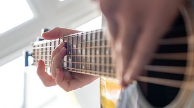 Akustik gitar çalan bir kişinin el görüntüsü, tellerin üzerine yerleştirilmiş parmaklar, bir pencereden gelen doğal ışıkla aydınlatılmış.