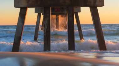 İskele yapısı, gün batımında düşen okyanus dalgalarının üzerinde yükselir. Islak kumu yansıtır ve renkli gökyüzü ile sakin bir sahne yaratır.