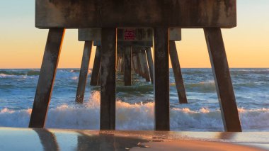 İskele yapısı, gün batımında düşen okyanus dalgalarının üzerinde yükselir. Islak kumu yansıtır ve renkli gökyüzü ile sakin bir sahne yaratır.