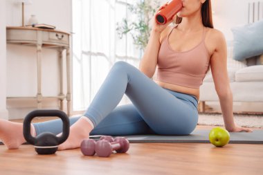 Formda bir kadın antrenmandan sonra dinleniyor. Kettlebell, su şişesi ve yeşil elma suyu ile yoga minderinde oturuyor. Sağlıklı yaşam tarzı, kişisel bakım ve iyileşme..
