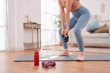 Evde kettlebell egzersiziyle beden eğitimi. Asyalı kadın yoga minderinde, güç topluyor ve rahat oturma odasında kaslarını geliştiriyor..
