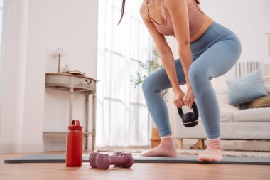 Evde kettlebell egzersiziyle beden eğitimi. Asyalı kadın yoga minderinde, güç topluyor ve rahat oturma odasında kaslarını geliştiriyor..