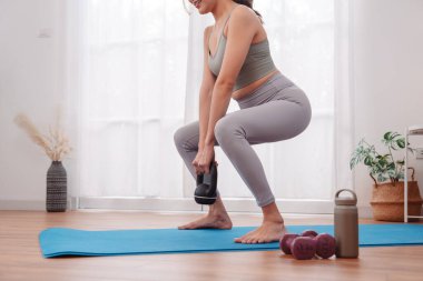 Squat exercise with kettlebell by Asian woman at home on yoga mat, strength training, lower body workout, and female fitness routine with minimal gym equipment.