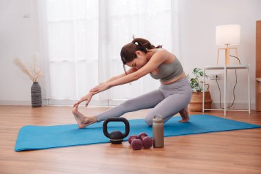 Evde yoga minderinde Asyalı bir kadınla egzersiz yapmak, ısınma rutini, kettlebell, dambıl ve spor salonu ve hareketlilik eğitimi için su şişesi..