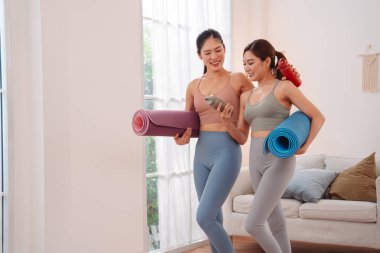 Young Asian women in activewear walking after yoga workout at home, holding yoga mats and water bottles. Fitness, wellness, exercise, healthy lifestyle, morning routine, stretching, selfcare.