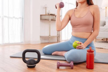 Formda bir kadın antrenmandan sonra dinleniyor. Kettlebell, su şişesi ve yeşil elma suyu ile yoga minderinde oturuyor. Sağlıklı yaşam tarzı, kişisel bakım ve iyileşme..