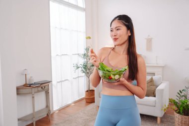 Aktif giyimli Asyalı kadın ve evde taze sebze salatası kasesi var. Temiz yeme, beslenme farkındalığı, sağlık alışkanlıkları ve dengeli bir yaşam tarzını teşvik eden ev konsepti sağlıklıdır..