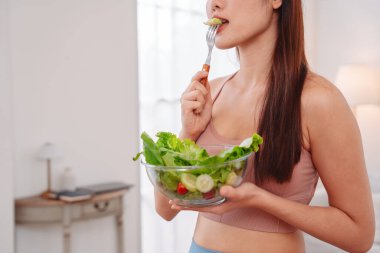 Aktif giyimli Asyalı kadın ve evde taze sebze salatası kasesi var. Temiz yeme, beslenme farkındalığı, sağlık alışkanlıkları ve dengeli bir yaşam tarzını teşvik eden ev konsepti sağlıklıdır..