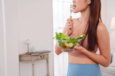 Aktif giyimli Asyalı kadın ve evde taze sebze salatası kasesi var. Temiz yeme, beslenme farkındalığı, sağlık alışkanlıkları ve dengeli bir yaşam tarzını teşvik eden ev konsepti sağlıklıdır..