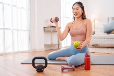 Kadın antremandan sonra mola veriyor, yoga minderinde halterler, kettlebell, yeşil elma ve su şişesiyle oturuyor. Sağlıklı yaşam tarzı, egzersiz sonrası iyileşme ve evdeki fitness beslenmesi..