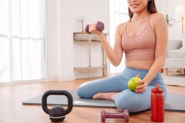 Kadın antremandan sonra mola veriyor, yoga minderinde halterler, kettlebell, yeşil elma ve su şişesiyle oturuyor. Sağlıklı yaşam tarzı, egzersiz sonrası iyileşme ve evdeki fitness beslenmesi..