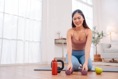Kadın antremandan sonra mola veriyor, yoga minderinde halterler, kettlebell, yeşil elma ve su şişesiyle oturuyor. Sağlıklı yaşam tarzı, egzersiz sonrası iyileşme ve evdeki fitness beslenmesi..