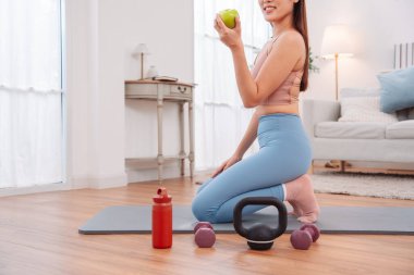 Kadın antremandan sonra mola veriyor, yoga minderinde halterler, kettlebell, yeşil elma ve su şişesiyle oturuyor. Sağlıklı yaşam tarzı, egzersiz sonrası iyileşme ve evdeki fitness beslenmesi..