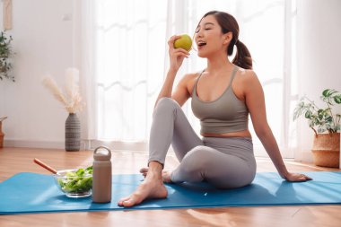 Genç Asyalı kadın, evdeki yoga minderinde egzersiz yaptıktan sonra dinleniyor, taze elma ve sağlıklı salatanın tadını çıkarıyor, sağlık alışkanlıklarını, dengeli beslenmeyi, aktif yaşam tarzını ve özenli egzersiz sonrası rahatlamayı vurguluyor..