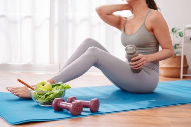 Genç Asyalı kadın, evdeki yoga minderinde egzersiz yaptıktan sonra dinleniyor, taze elma ve sağlıklı salatanın tadını çıkarıyor, sağlık alışkanlıklarını, dengeli beslenmeyi, aktif yaşam tarzını ve özenli egzersiz sonrası rahatlamayı vurguluyor..