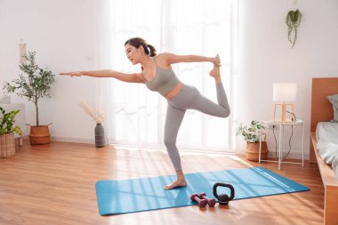 Genç Asyalı kadın yatak odasındaki minder üzerinde yoga yapıyor, tek bacağının üzerinde odaklanmış bir ifadeyle dengede duruyor. Sağlıklı yaşam tarzı, esneklik, farkındalık ve sakin bir yerde sağIık rutini..