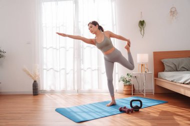 Genç Asyalı kadın yatak odasındaki minder üzerinde yoga yapıyor, tek bacağının üzerinde odaklanmış bir ifadeyle dengede duruyor. Sağlıklı yaşam tarzı, esneklik, farkındalık ve sakin bir yerde sağIık rutini..