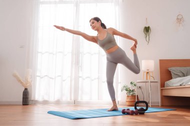 Genç Asyalı kadın yatak odasındaki minder üzerinde yoga yapıyor, tek bacağının üzerinde odaklanmış bir ifadeyle dengede duruyor. Sağlıklı yaşam tarzı, esneklik, farkındalık ve sakin bir yerde sağIık rutini..