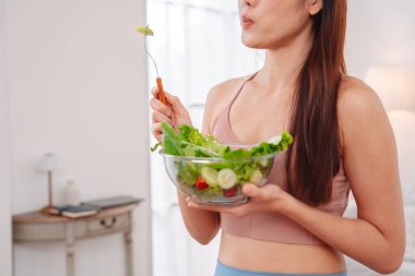 Aktif giyimli Asyalı kadın ve evde taze sebze salatası kasesi var. Temiz yeme, beslenme farkındalığı, sağlık alışkanlıkları ve dengeli bir yaşam tarzını teşvik eden ev konsepti sağlıklıdır..