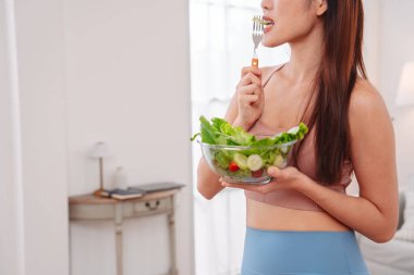 Aktif giyimli Asyalı kadın ve evde taze sebze salatası kasesi var. Temiz yeme, beslenme farkındalığı, sağlık alışkanlıkları ve dengeli bir yaşam tarzını teşvik eden ev konsepti sağlıklıdır..